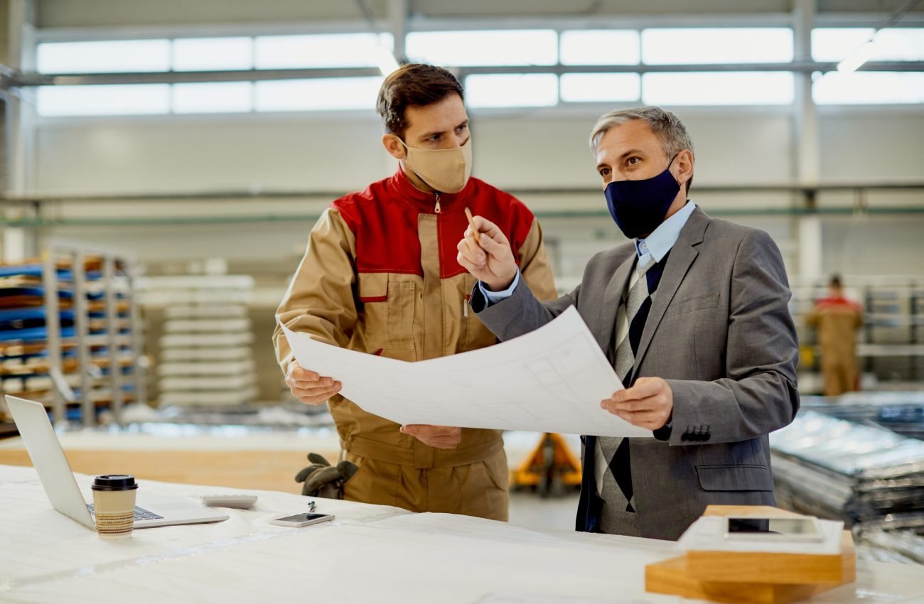 Mid adult businessman and manual worker analyzing plans while working at carpentry workshop during coronavirus pandemic.